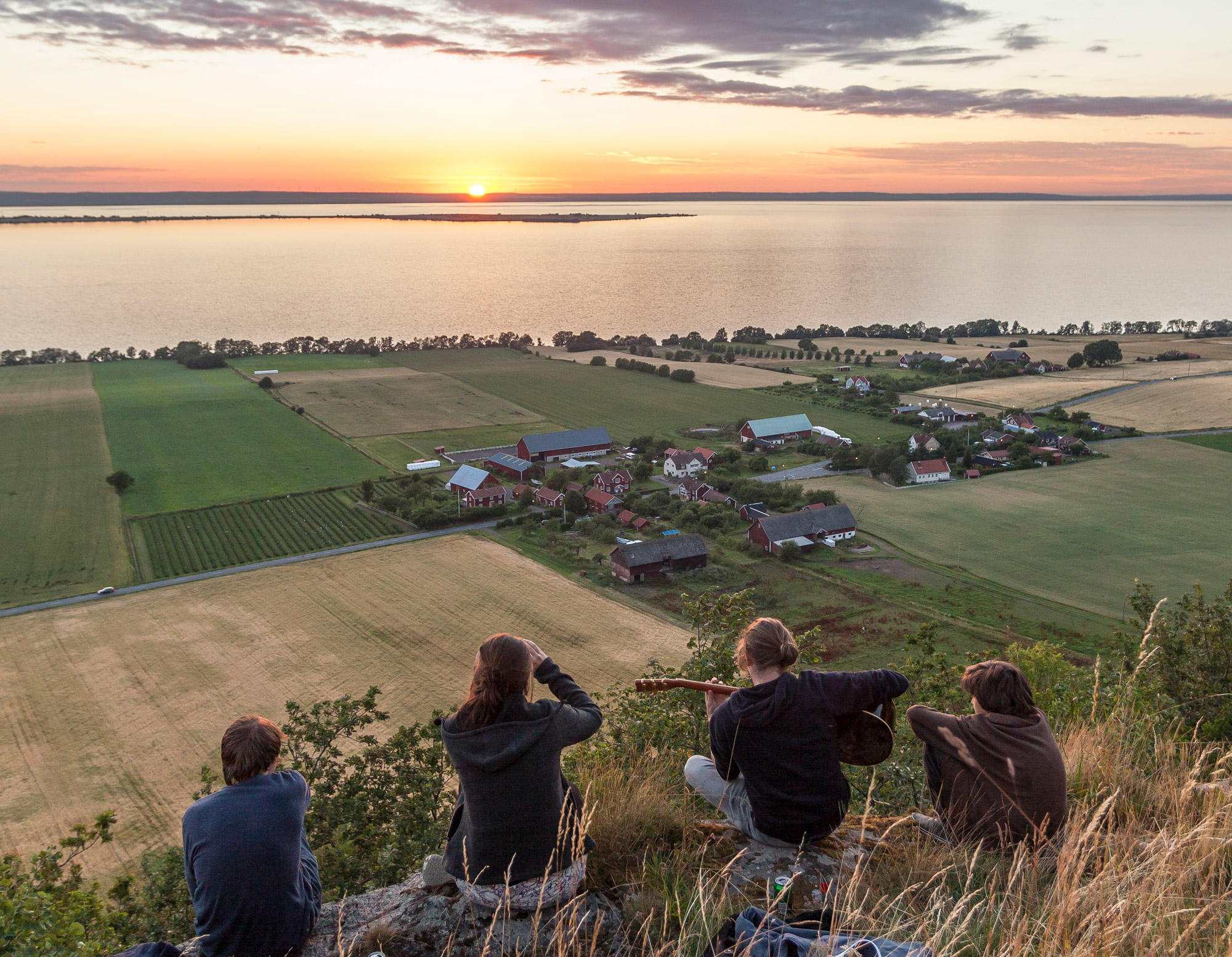 Ett kompisgäng med gitarr sitter på baksidan av Brahehus och tittar på solnedgången över Visingsö