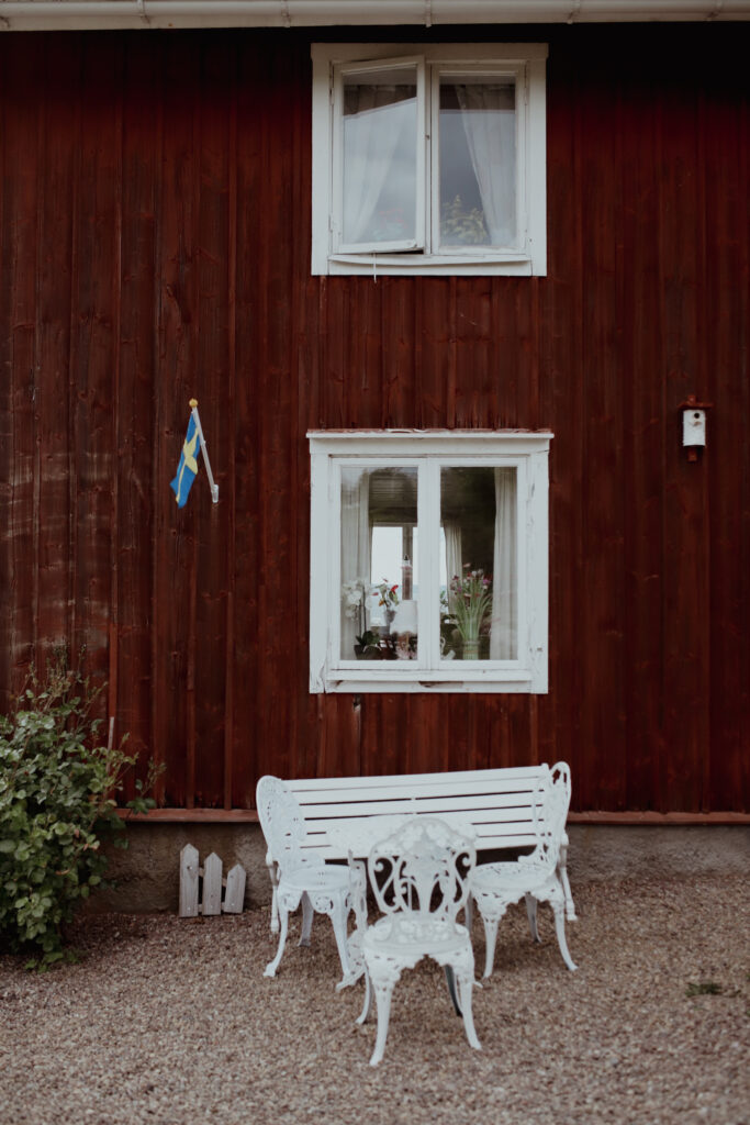 En röd husfasad med vita fönster. Utanför står en vit bänk med vita stolar.