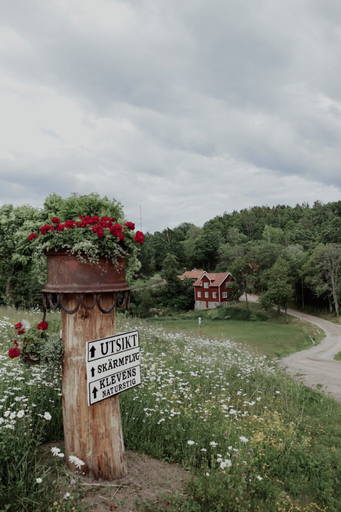 Skylt med blomma på stolpen. I bakgrunden ett rött hus med vita knutar.